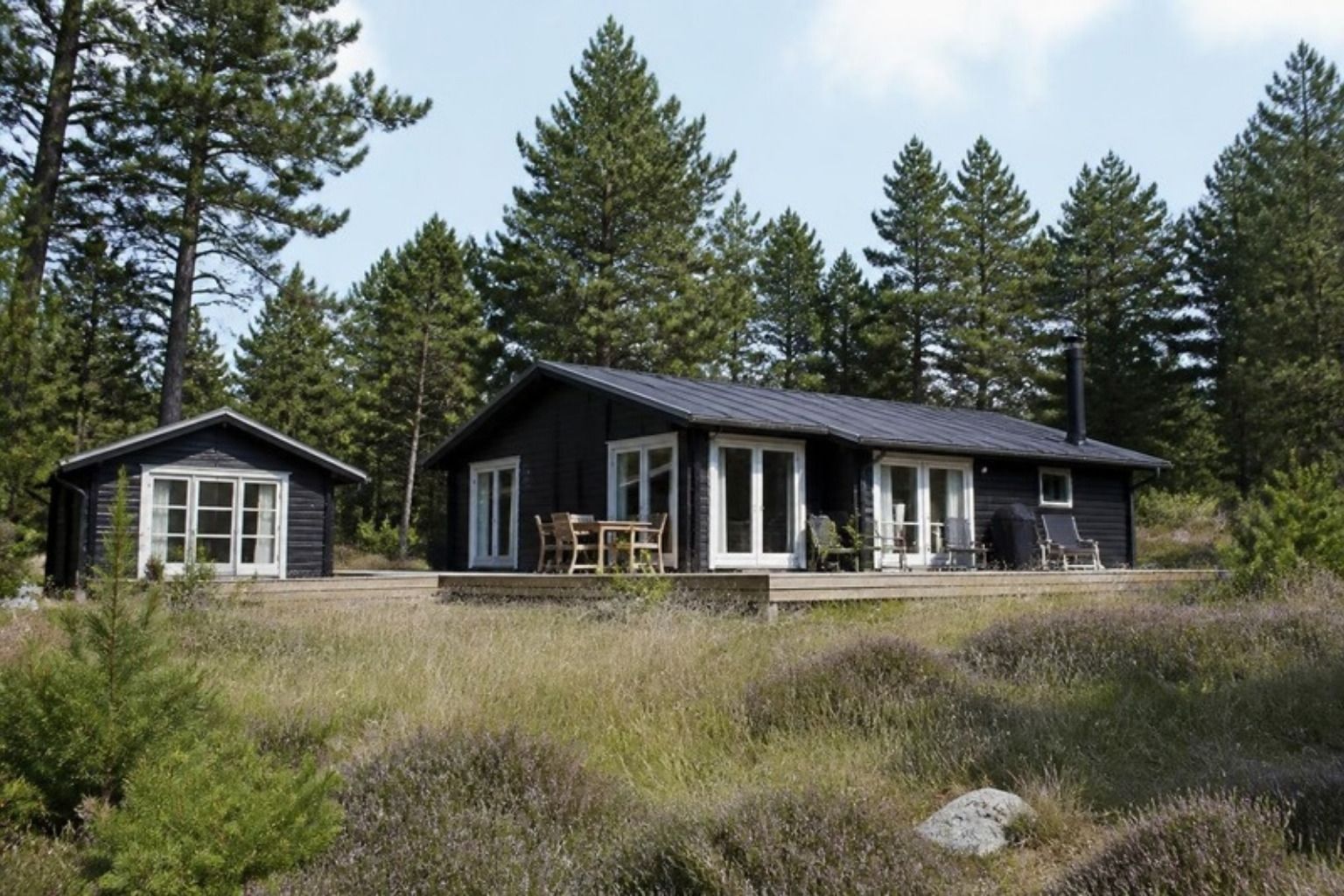 Sort sommerhus og anneks på en grund med lyng og fyrretræer