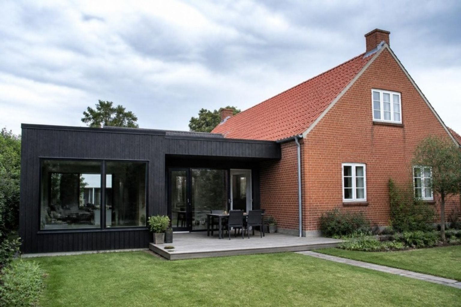 Sort tilbygning i træ med store vinduer sat sammen med et rødt murstenshus